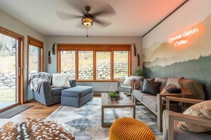 Ein modernes Wohnzimmer mit einem grauen Sofa, einem blauen Sessel und einem Holztisch. Ein großes Fenster bietet Blick auf eine Steinsäule und ein leuchtendes Neonzeichen an der Wand.