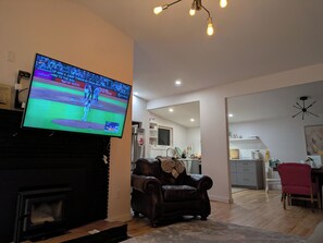 A living room with a flat-screen TV, a fireplace, a leather armchair, and a dining area with a red chair and a wooden table.
