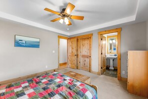 Une chambre à coucher avec un lit, un ventilateur de plafond, une porte en bois et une salle de bain visible à travers une porte ouverte.