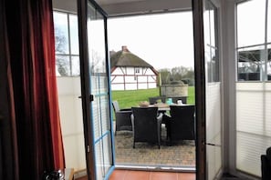 Eine geöffnete Tür führt zu einer Terrasse mit Tisch und Stühlen, im Hintergrund ein Haus mit Strohdach und eine Rasenfläche.