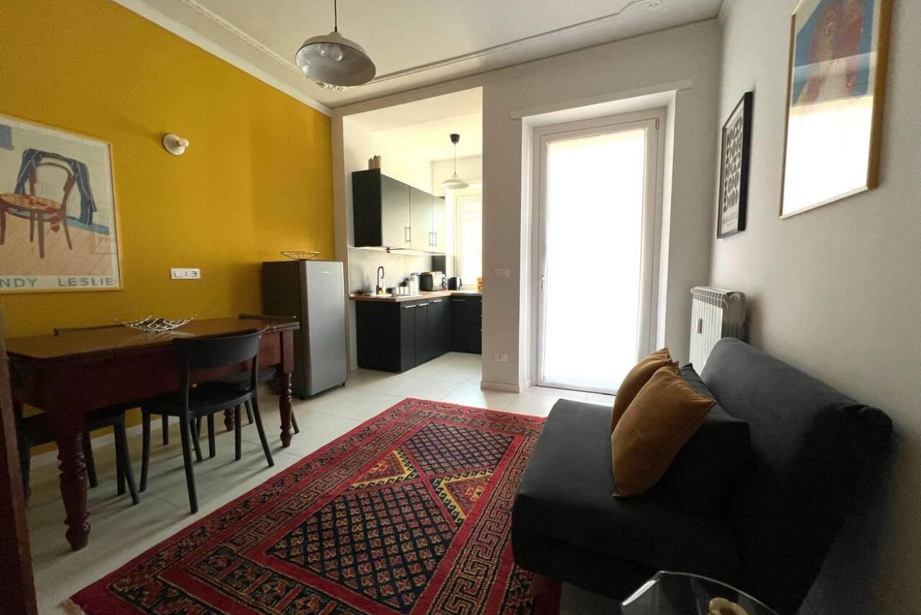 Un ambiente compatto con angolo ristoro, divano e cucina sullo sfondo.