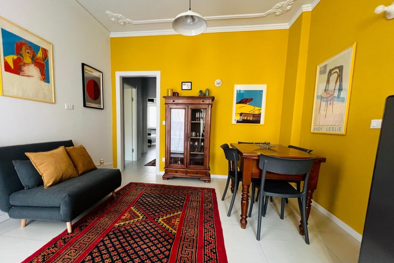Ein Wohnzimmer mit einer Couch, einem Esstisch mit Stühlen, einem gemusterten Teppich und Bildern an den Wänden.