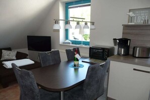 Ein kompakter Wohnbereich mit Esstisch, Stühlen, einem Flachbildfernseher, einem Fenster mit Aussicht und einer Küchenzeile, inklusive Kaffeemaschine und Toaster.