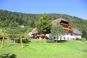 A two-story house with a sloped roof, surrounded by a well-maintained lawn and a forested hill in the background. - Holiday apartment with 2 bedrooms (Oppenau)