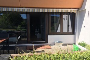 A building with a sliding glass door entrance, a small outdoor seating area with a table and chairs, and a green watering can on the ground. - Holiday apartment with dog in Dahn (Dahn)
