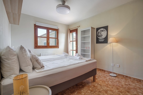 Un dormitorio con cama, radiador, ventana, puerta, estante montado en la pared, lámpara de pie y cuadro colgado.