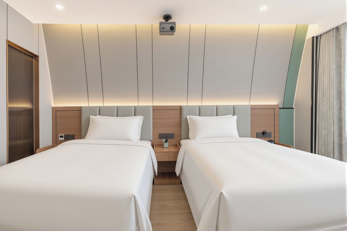 Habitación de hotel con dos camas, un cabecero de madera, una mesita de noche con una cámara y una ventana con cortinas.