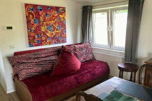 Ein Zimmer mit einer Couch, einem Tisch, einem Sessel, einem Fenster mit Vorhängen und einem farbenfrohen abstrakten Gemälde an der Wand.