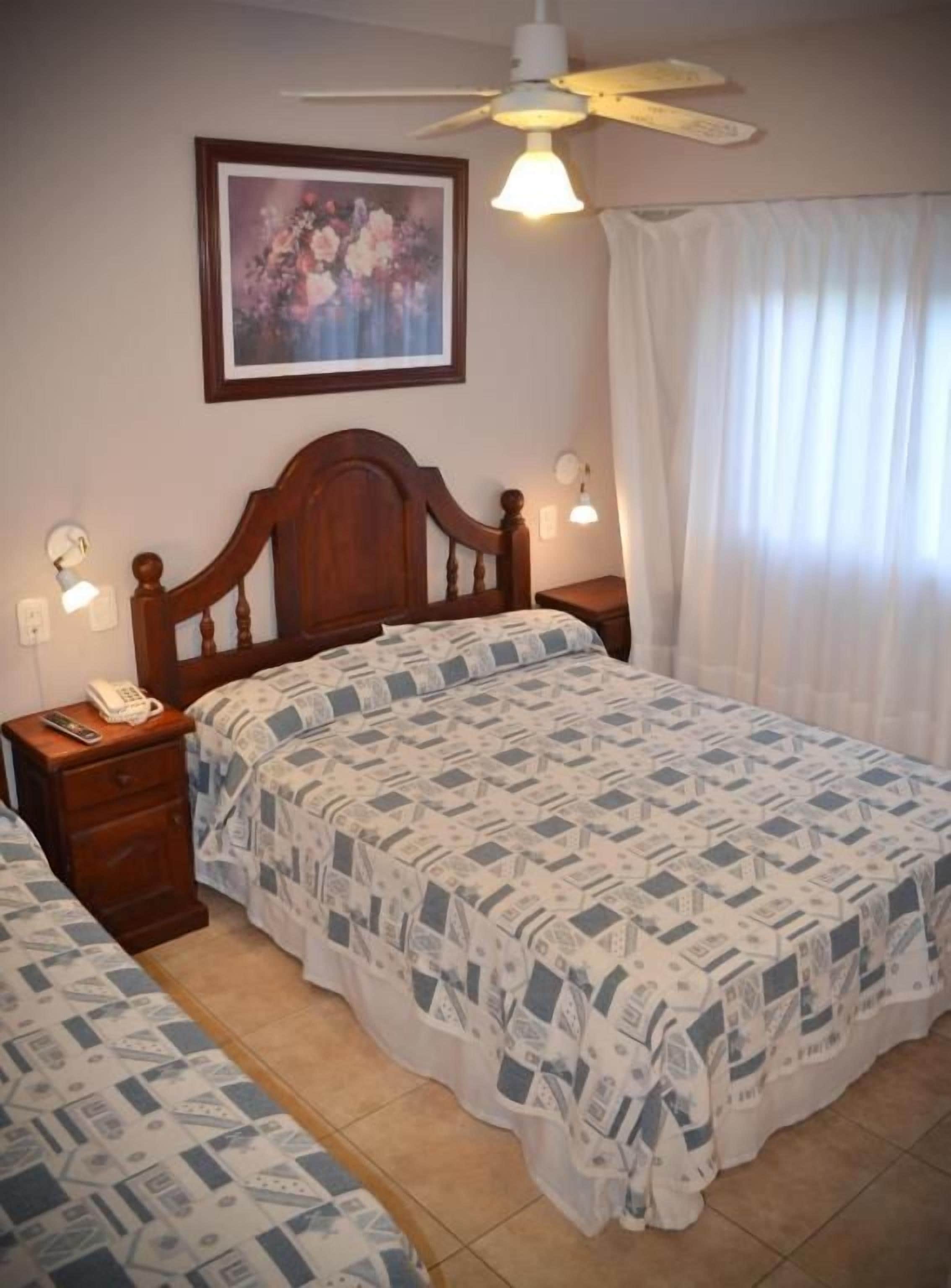 A bedroom with a wooden bed, a ceiling fan, a bedside table with a lamp, and a framed picture on the wall.
