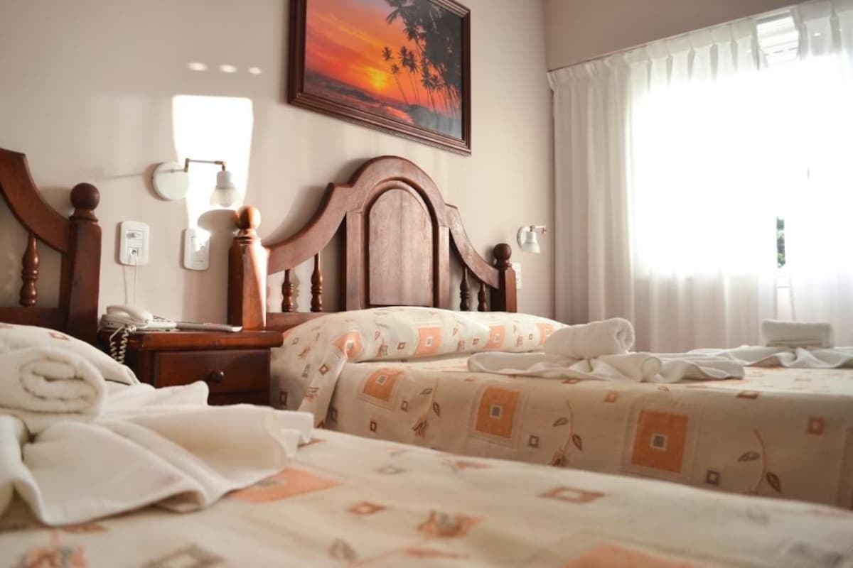 A hotel room with a wooden headboard, a bedside table with a lamp, a telephone, and a painting of a sunset.