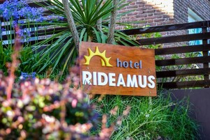 Un letrero de madera con la palabra 'hotel' y 'RIDEAMUS', ubicado frente a una cerca con follaje.