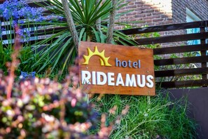A wooden sign with the word 'hotel' and 'RIDEAMUS' on it, placed in front of a fence with greenery. - Hotel Rideamus (Villa Gesell)