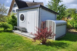 A small, single-story building with a circular window and a door, surrounded by a well-maintained lawn and a small tree with red leaves. - Holiday apartment Bug (Dranske)