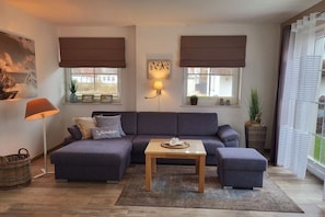 A living room with a sofa, ottoman, and coffee table. There are two windows with blinds, a lamp, and a wall-mounted light. - Holiday apartment Bug (Dranske)
