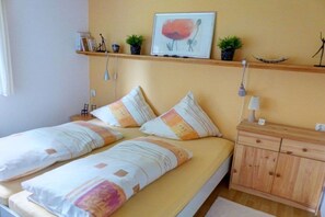 A double bed with striped bedding, a bedside table with a lamp, a wooden nightstand, and a shelf with decorative items. - Holiday home with 1 bedroom (Marienberg\/OT Rittersberg)