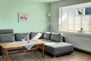 Ein Zimmer mit grauem Sofa, Holztisch, Stehlampe und einem Fenster mit Jalousien.