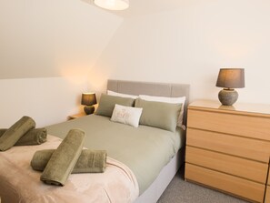 Ein ordentlich bezogenes Bett mit Kissen und Decke, eine Nachttischlampe, eine Holzkommode und eine Wandleuchte.