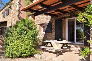 Ein Steinhaus mit überdachter Terrasse, auf der ein Picknicktisch steht, und einem Glastür, die zu einem Innenbereich führt.