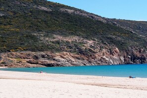 Ein Strand mit klarem, blauem Wasser, sandigem Ufer und einer hügeligen, felsigen Küste im Hintergrund.