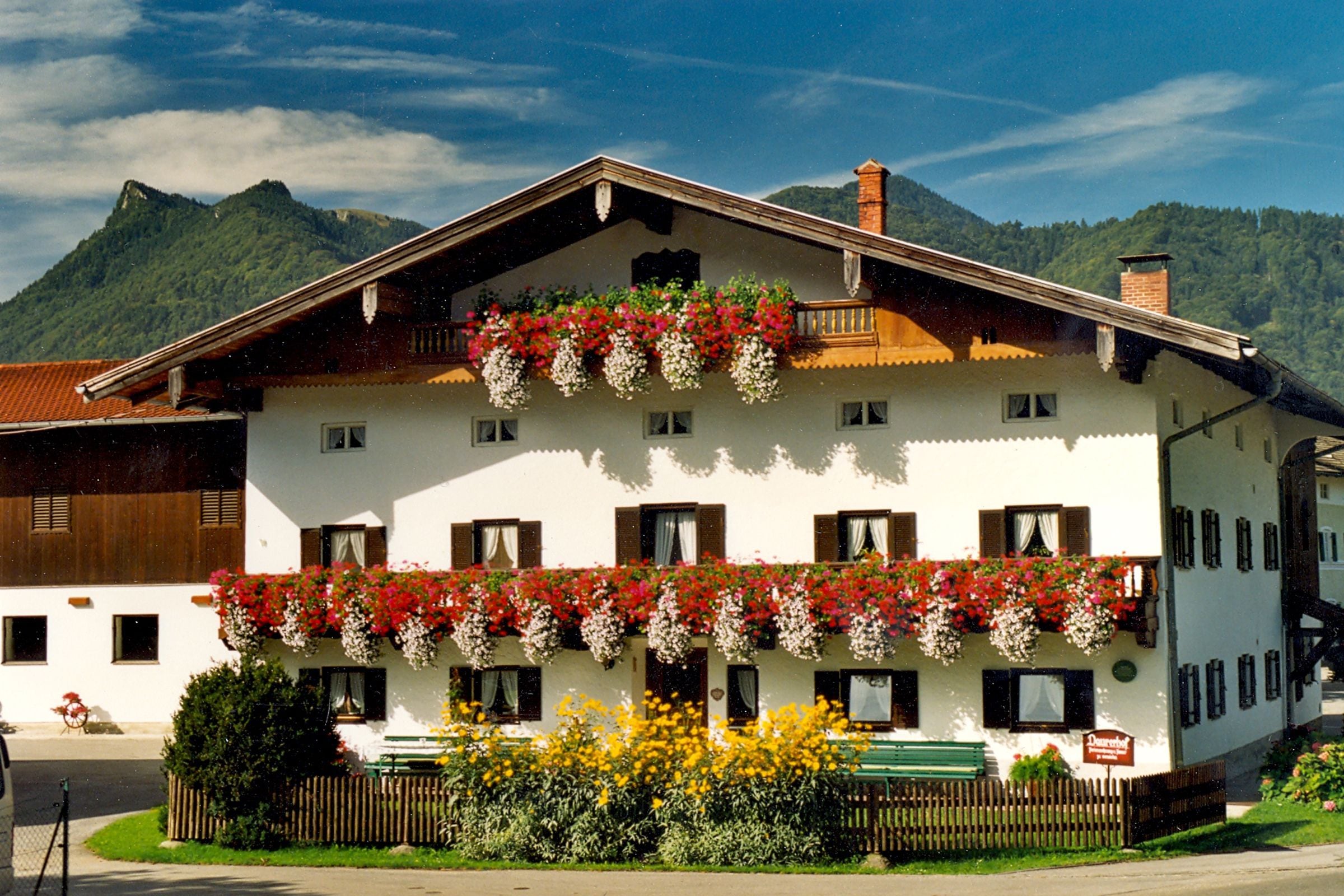 Un edificio de dos pisos con un balcón decorado con flores, una valla de madera y montañas al fondo.