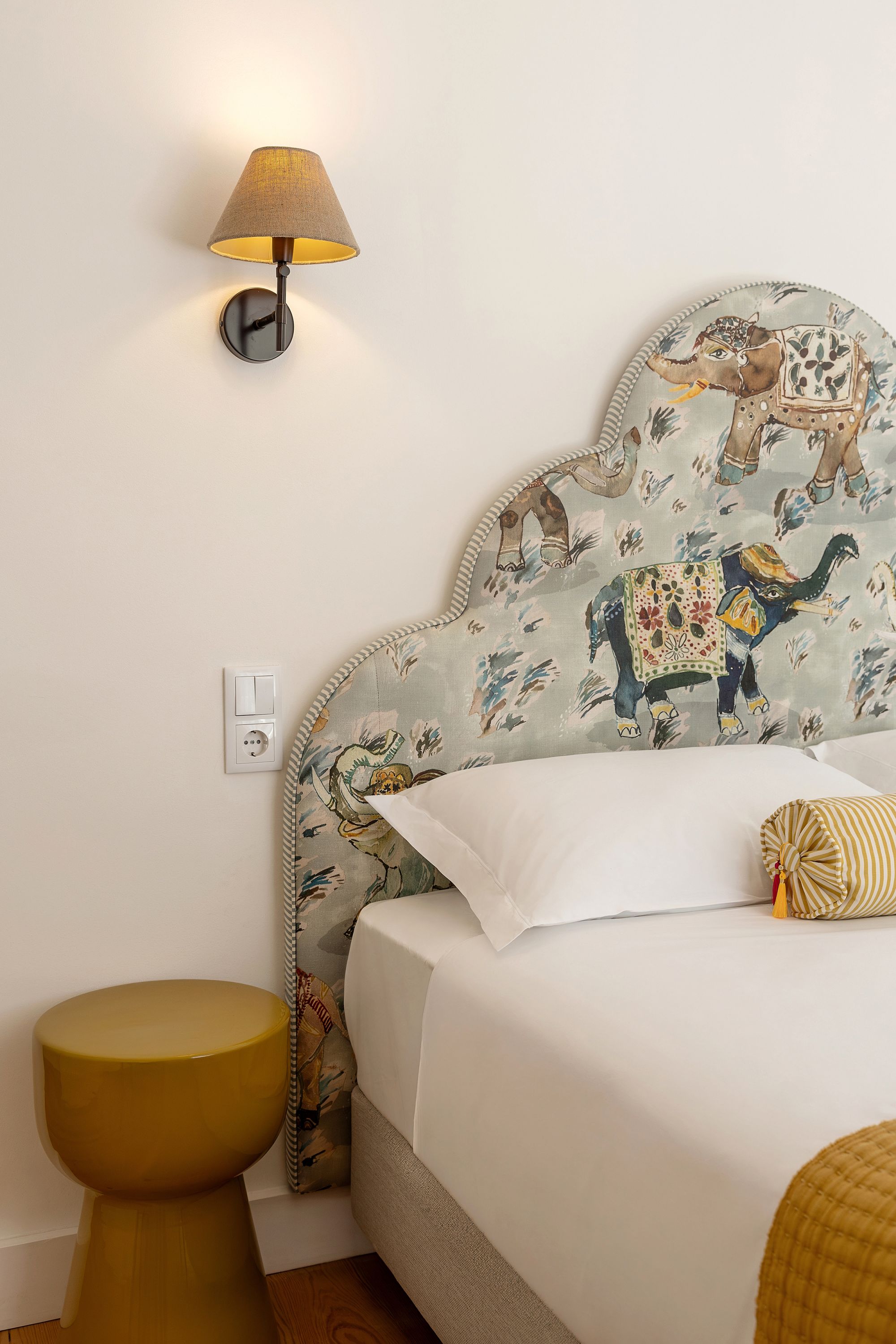 Ein Schlafzimmer mit einem gemusterten Kopfteil, einer Nachttischlampe, einem weißen Bett und einem gelben Hocker.