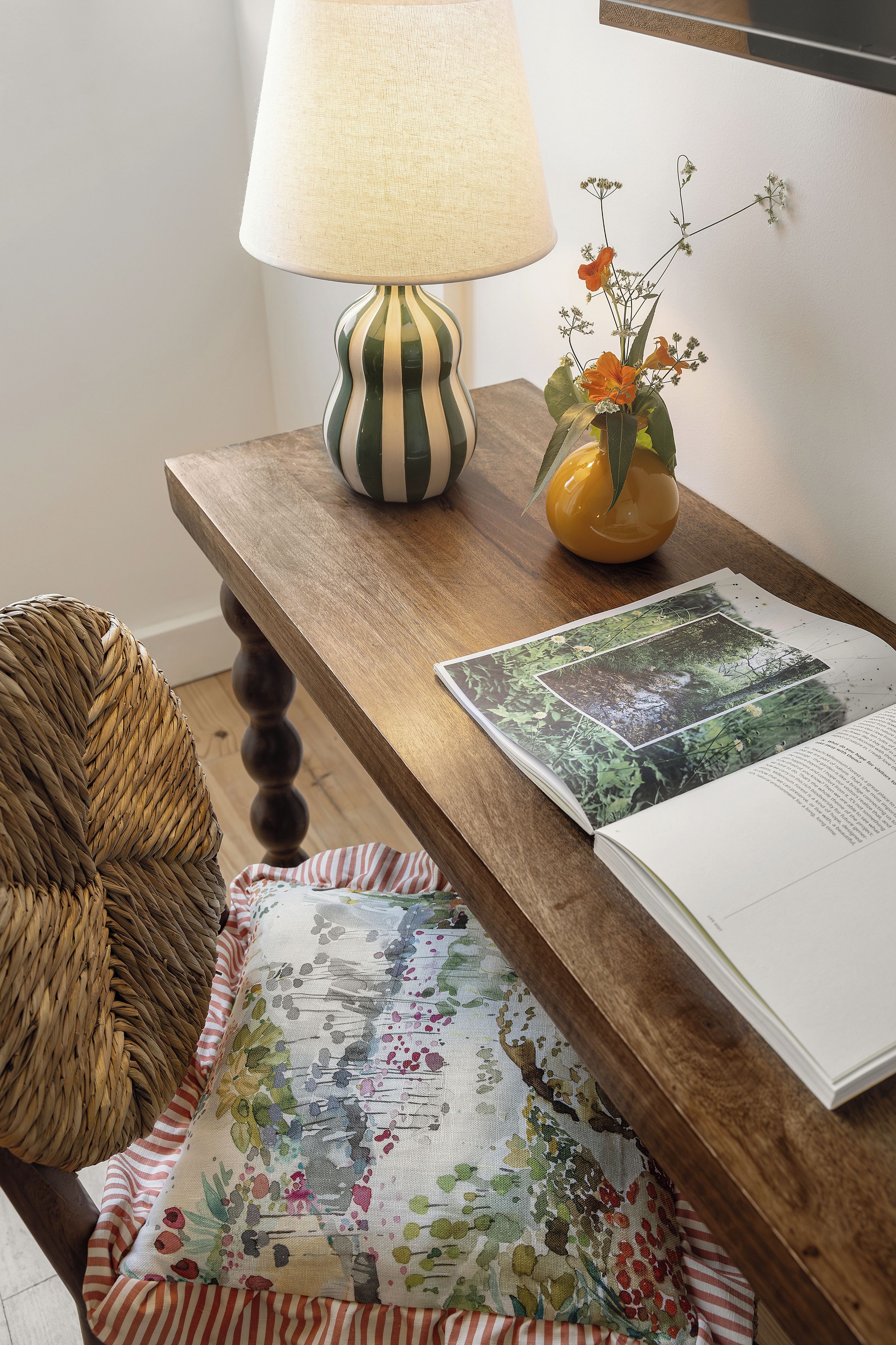 Ein Holztisch mit einer Lampe, einer Blumenvase, einem gewebten Korb und einem Buch mit einem Motiv aus der Natur.