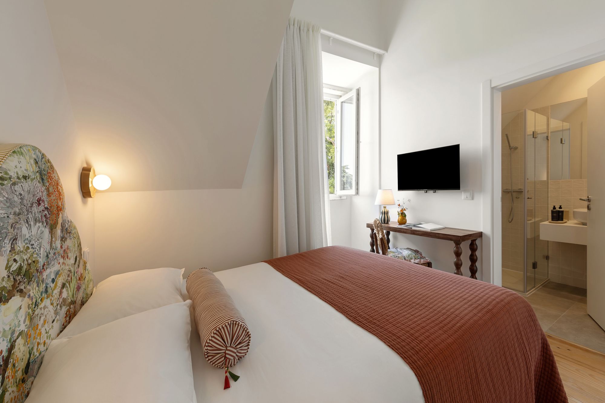 A hotel room with a bed, a television, a desk with a lamp, and a bathroom visible through an open door.