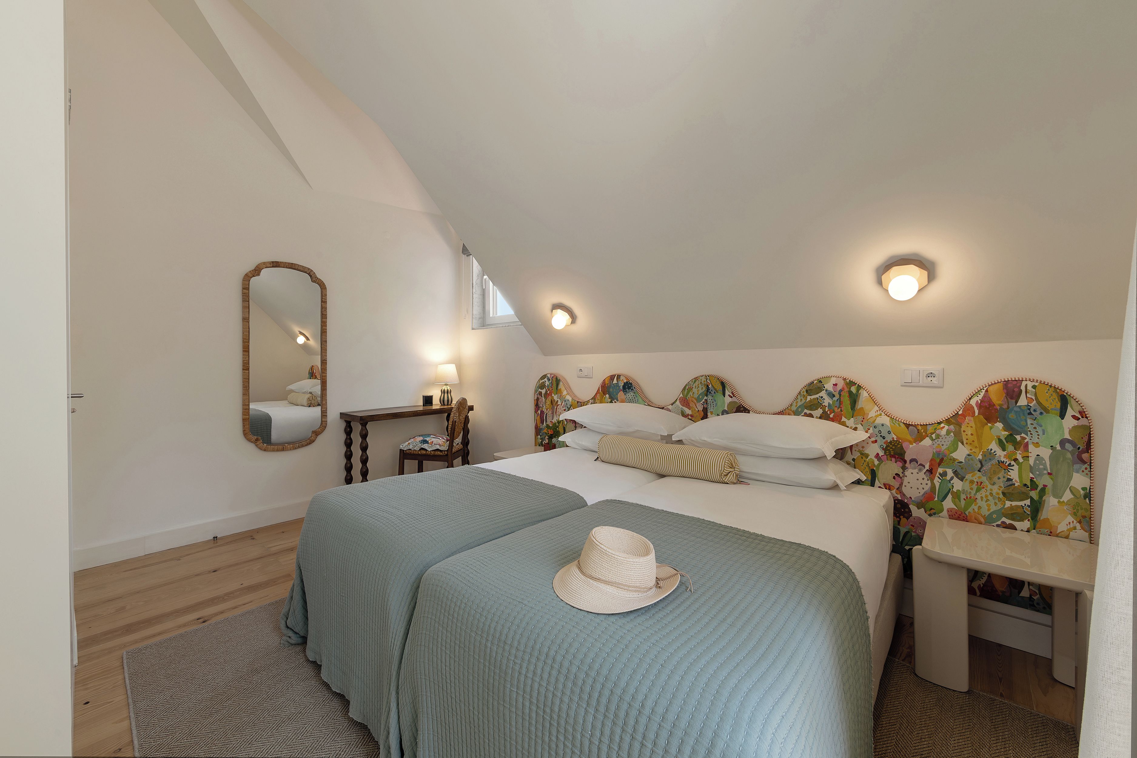 A bedroom with a sloped ceiling, a bed with two pillows, a bedside table, a chair, a mirror, and a window.