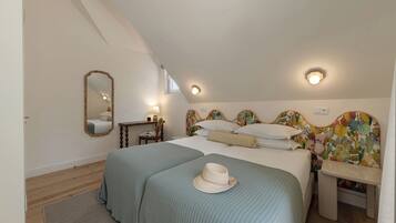 A bedroom with a sloped ceiling, a bed with two pillows, a bedside table, a chair, a mirror, and a window.