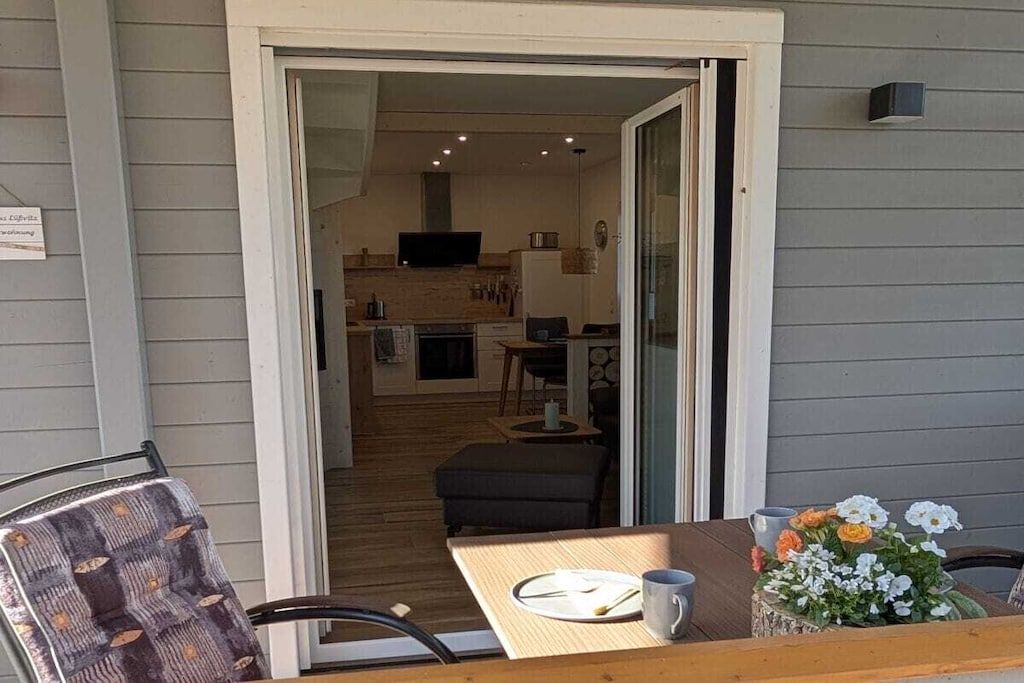 A room with a dining table, chairs, a small kitchen area, and a television.