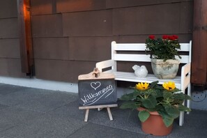 Eine hölzerne Bank mit einem Kreidetafel-Schild, ein Blumentopf mit roten Blüten und ein Blumentopf mit gelben Blüten.