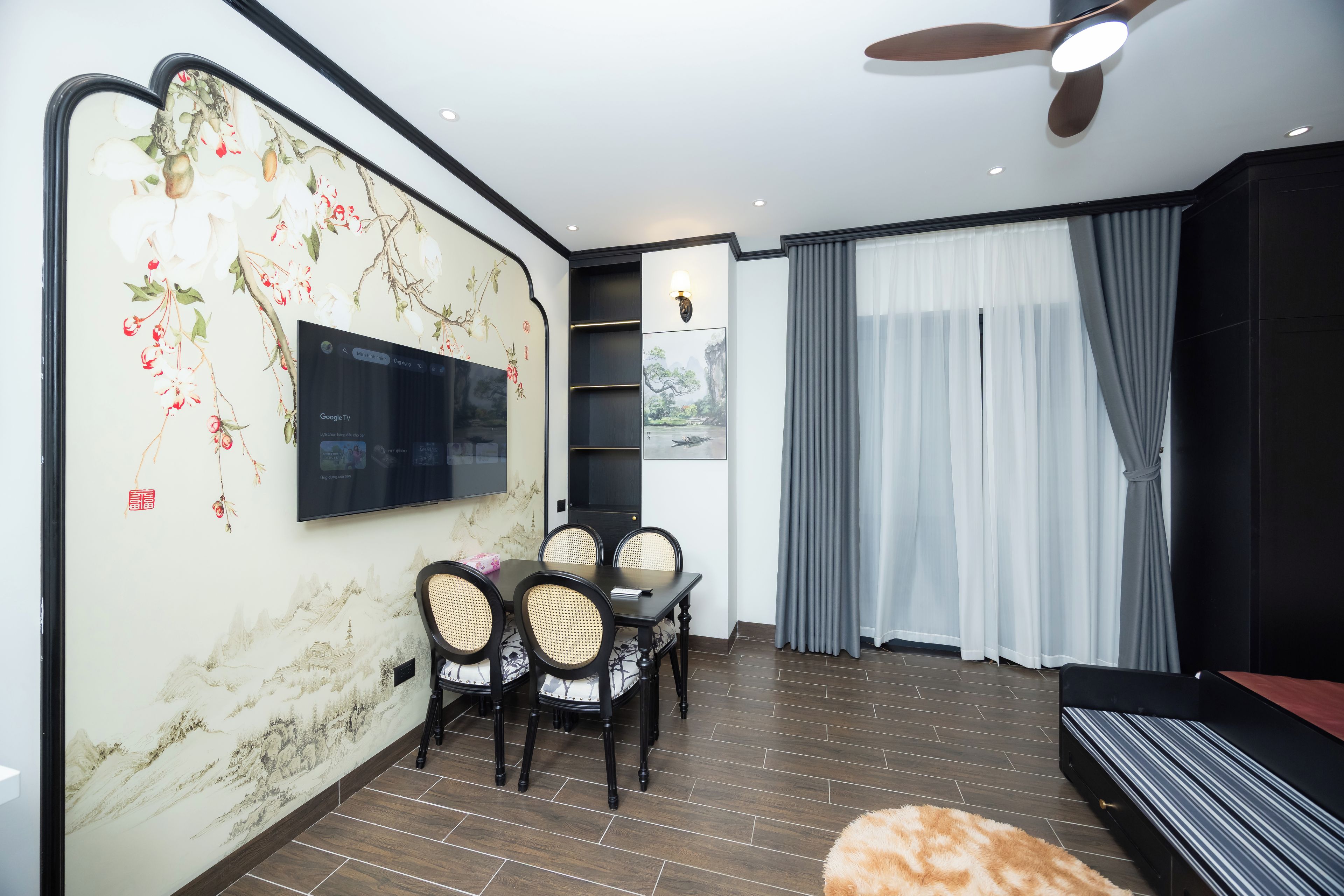 A modern living room with a flat-screen TV mounted on a wall with floral wallpaper, a dining area with a table and chairs, a ceiling fan, and a large window with curtains.
