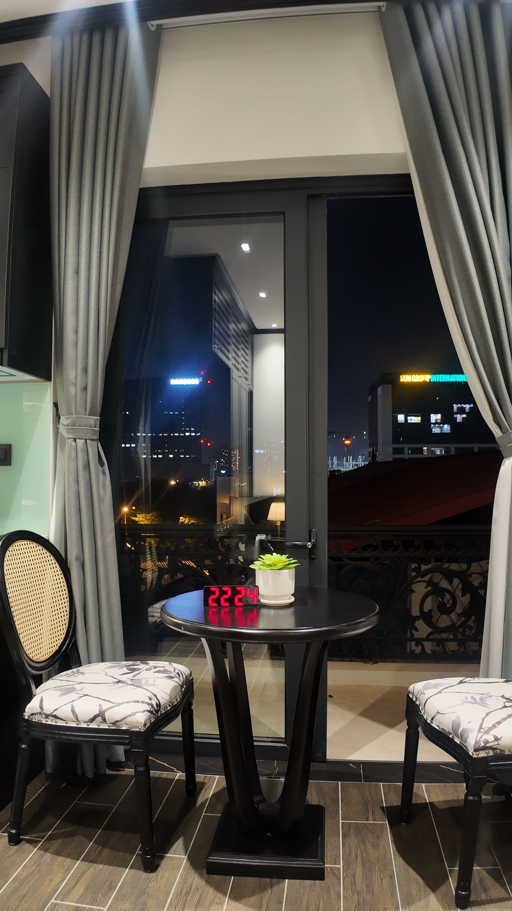 Un balcone con un tavolo, due sedie, una pianta in vaso e una sveglia digitale che mostra l'ora.