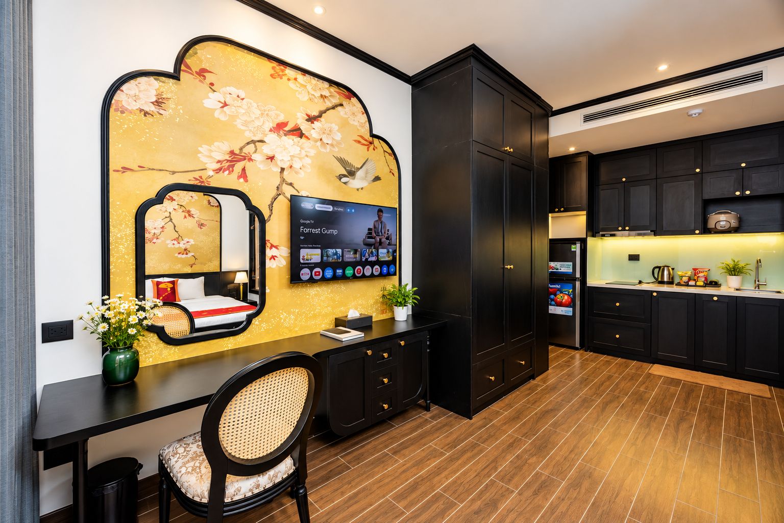 Ein modernes Hotelzimmer mit einem großen Fernseher, einem Schreibtisch mit Stuhl, einer Wand mit floralem Dekor und einer Küchenzeile mit dunklen Schränken und einer Arbeitsplatte.