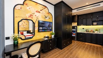 Ein modernes Hotelzimmer mit einem großen Fernseher, einem Schreibtisch mit Stuhl, einer Wand mit floralem Dekor und einer Küchenzeile mit dunklen Schränken und einer Arbeitsplatte.
