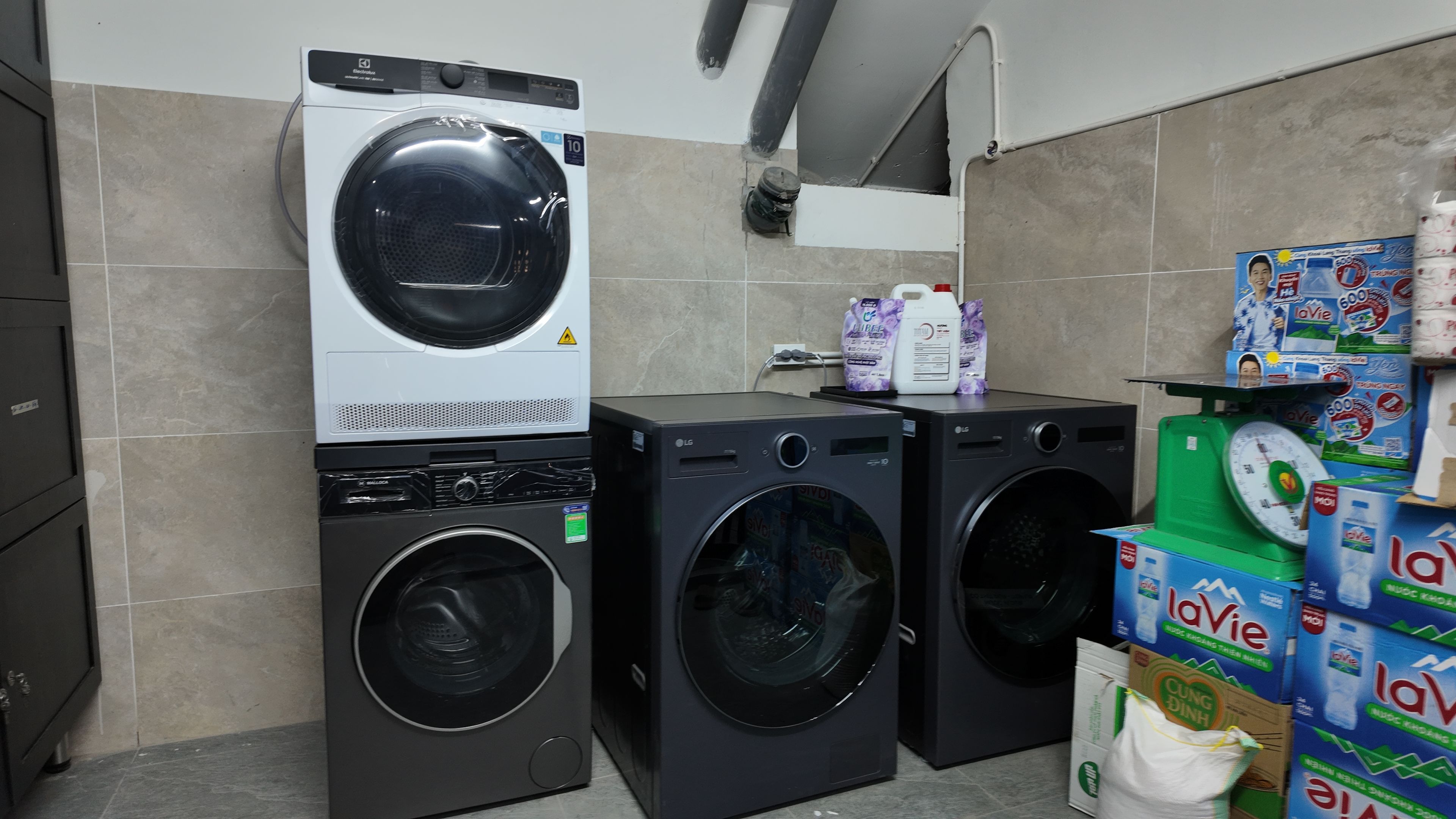 A laundry room with two stacked washing machines, a scale, and detergent boxes.