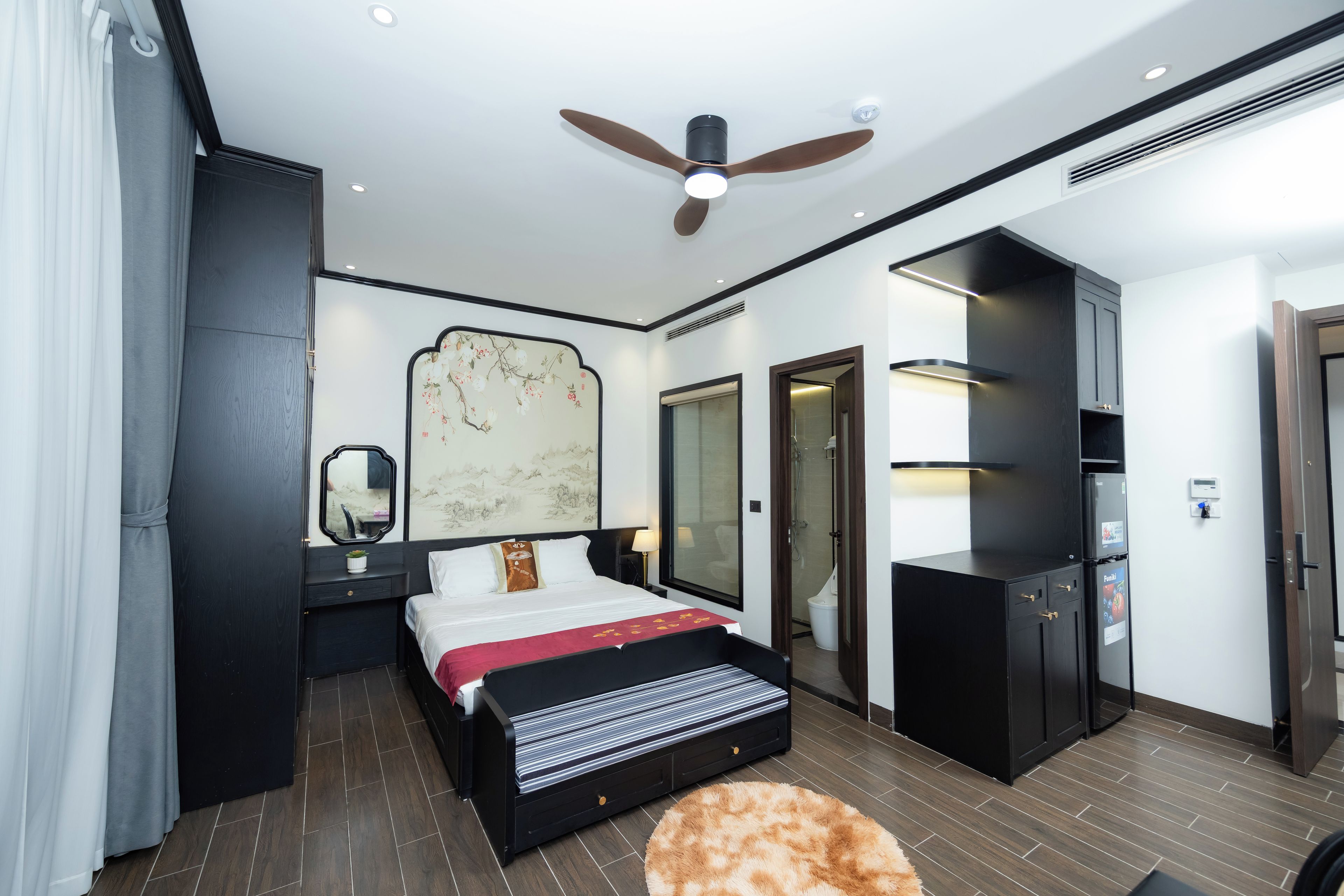 A modern hotel room with a bed, a ceiling fan, a large mirror, a wall-mounted TV, and a wardrobe.