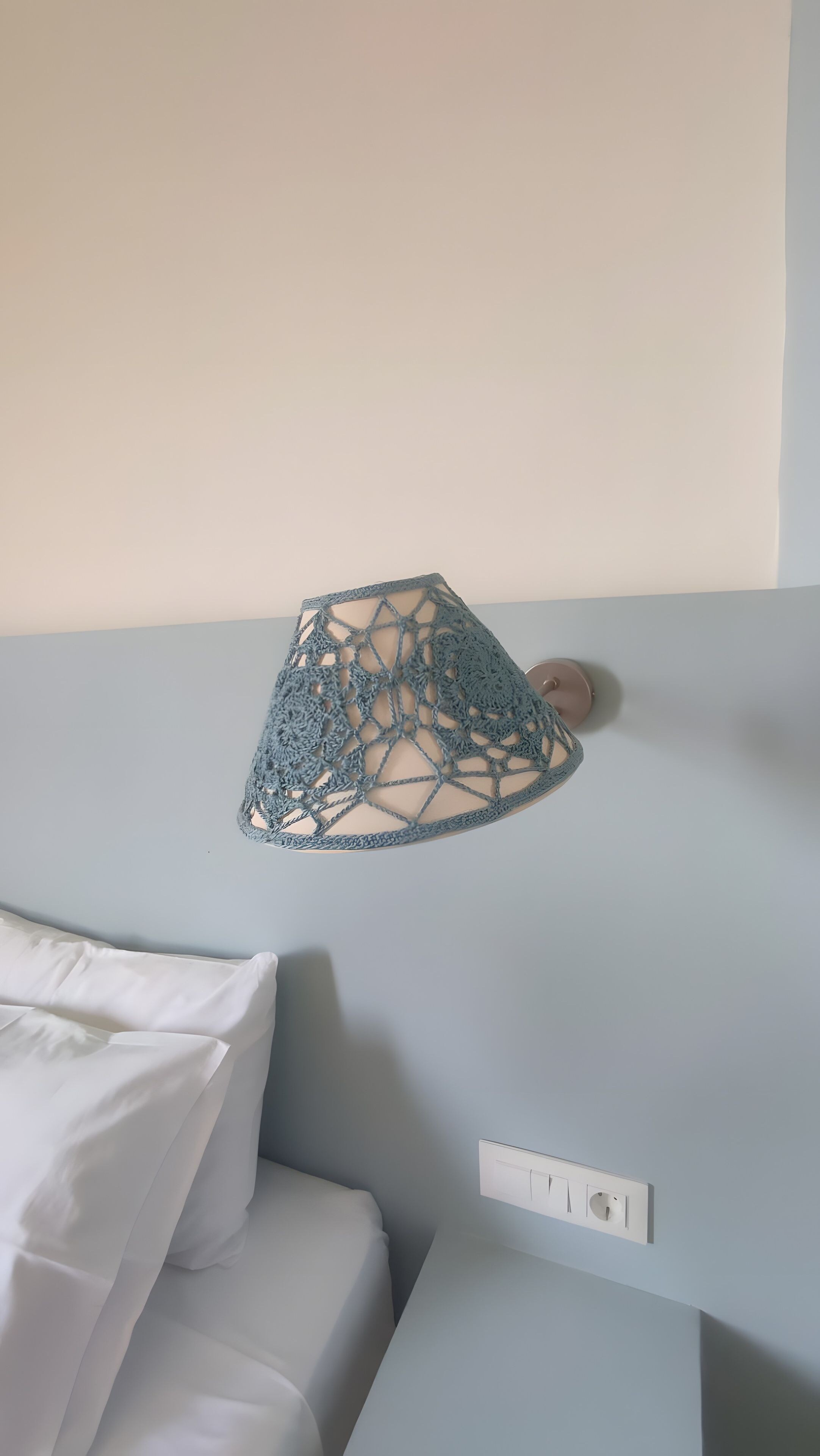 A bed with white bedding and a blue crocheted lampshade on a wall-mounted light fixture.