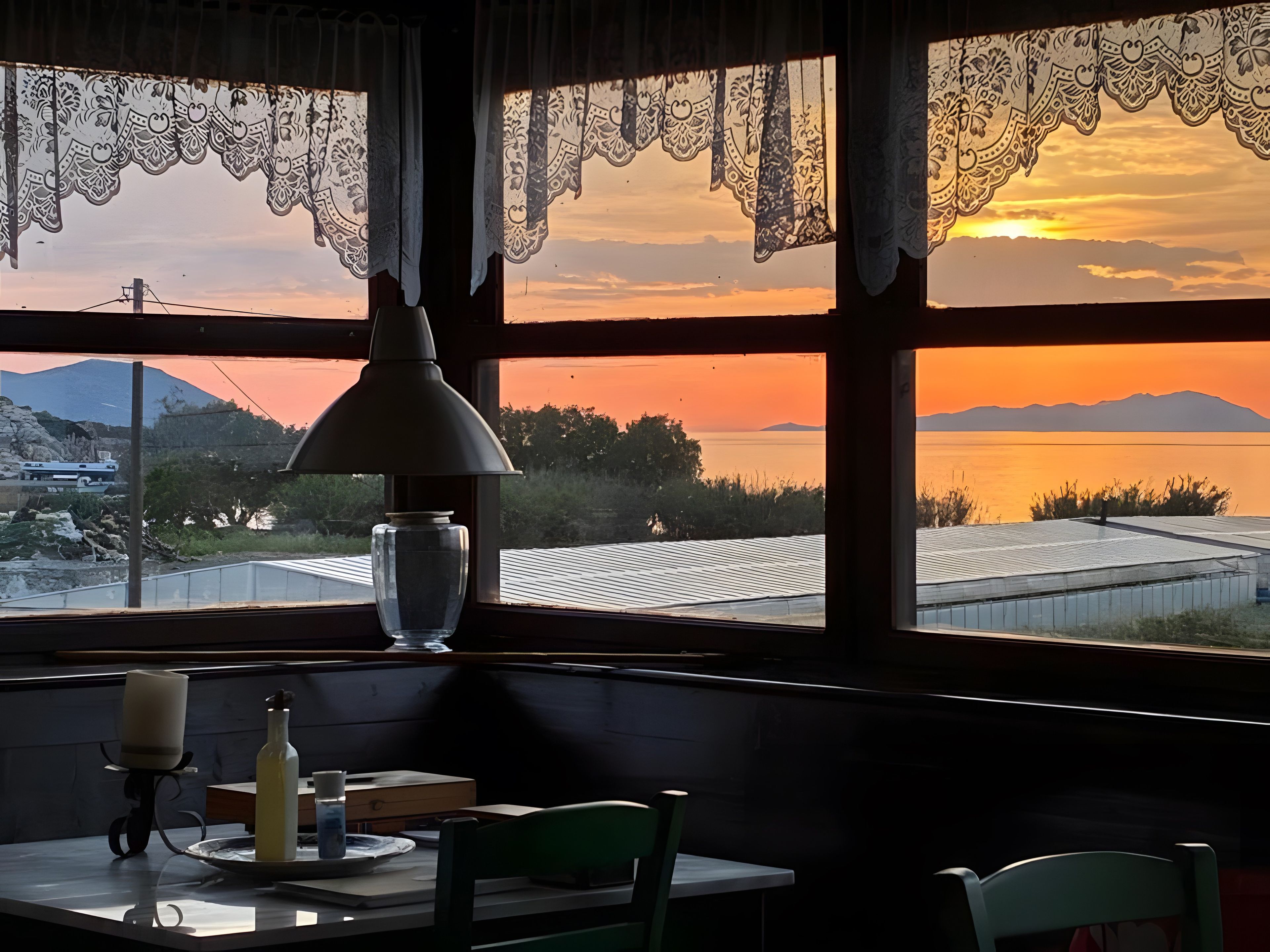 A table with a candle, a bottle, and a book, a window with lace curtains, and a view of a sunset over water and mountains.