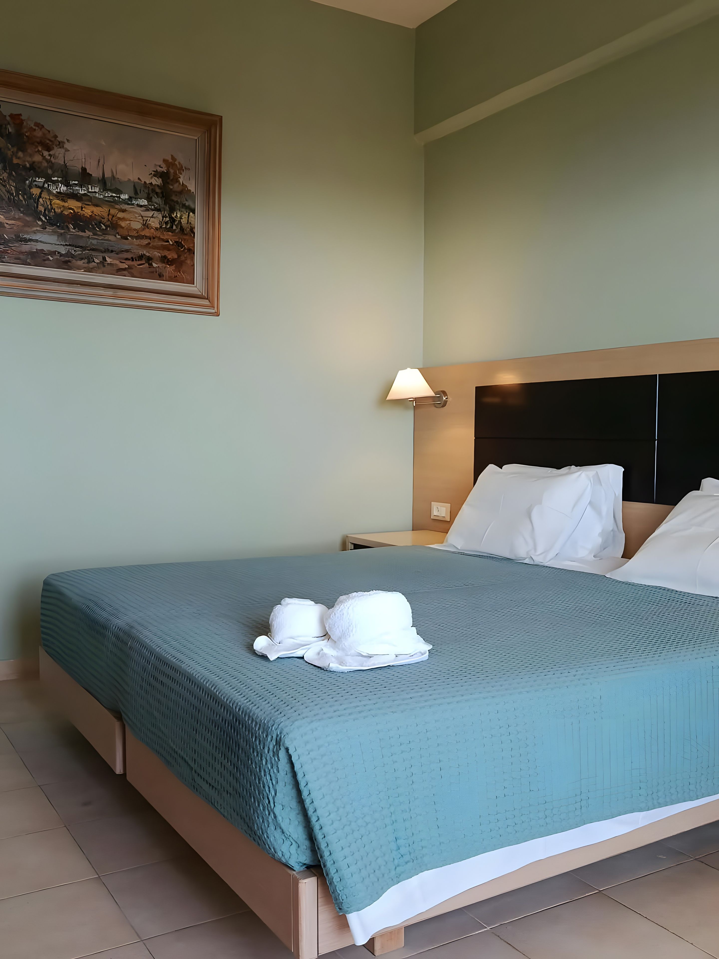 A neatly made bed with a teal bedspread and white pillows, a nightstand with a lamp, and a framed landscape painting on the wall.