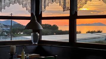 Une table avec une bougie, une bouteille et un livre, une fenêtre ornée de rideaux en dentelle, et une vue sur un coucher de soleil se reflétant sur l’eau et les montagnes.