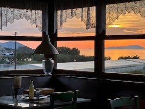 Una mesa con una vela, una botella y un libro, una ventana con cortinas de encaje, y una vista del atardecer sobre el agua y las montañas.