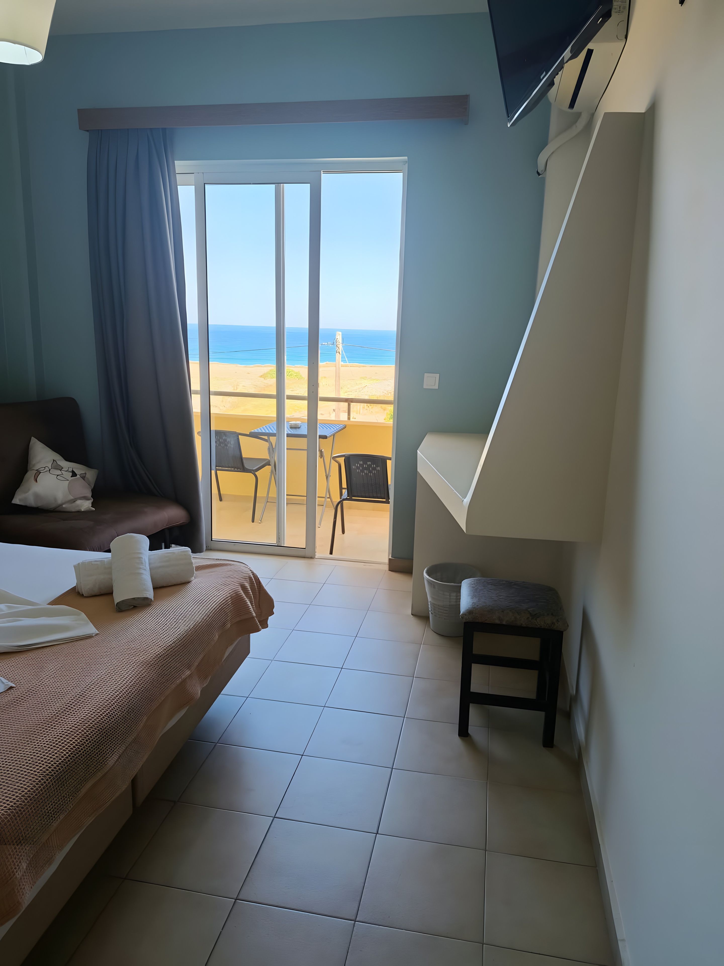 A hotel room with a bed, a small table, a chair, a TV, and a view of the beach.