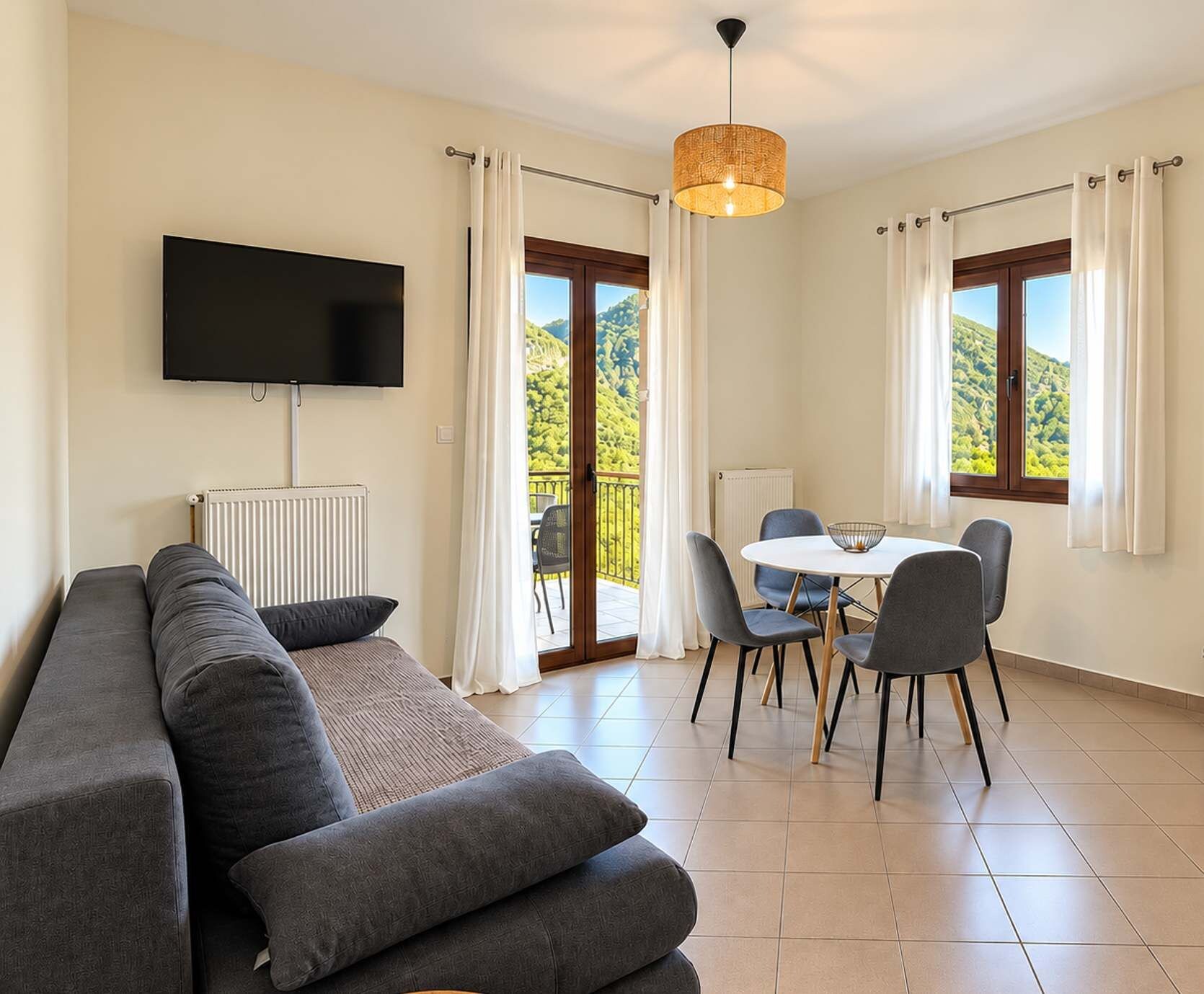 A modern living room with a sofa, a flat-screen TV, a dining table with chairs, and a view of a mountainous landscape through large windows.