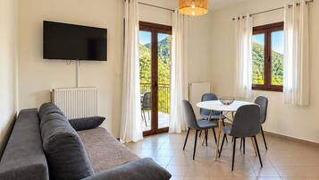 A modern living room with a sofa, a flat-screen TV, a dining table with chairs, and a view of a mountainous landscape through large windows.