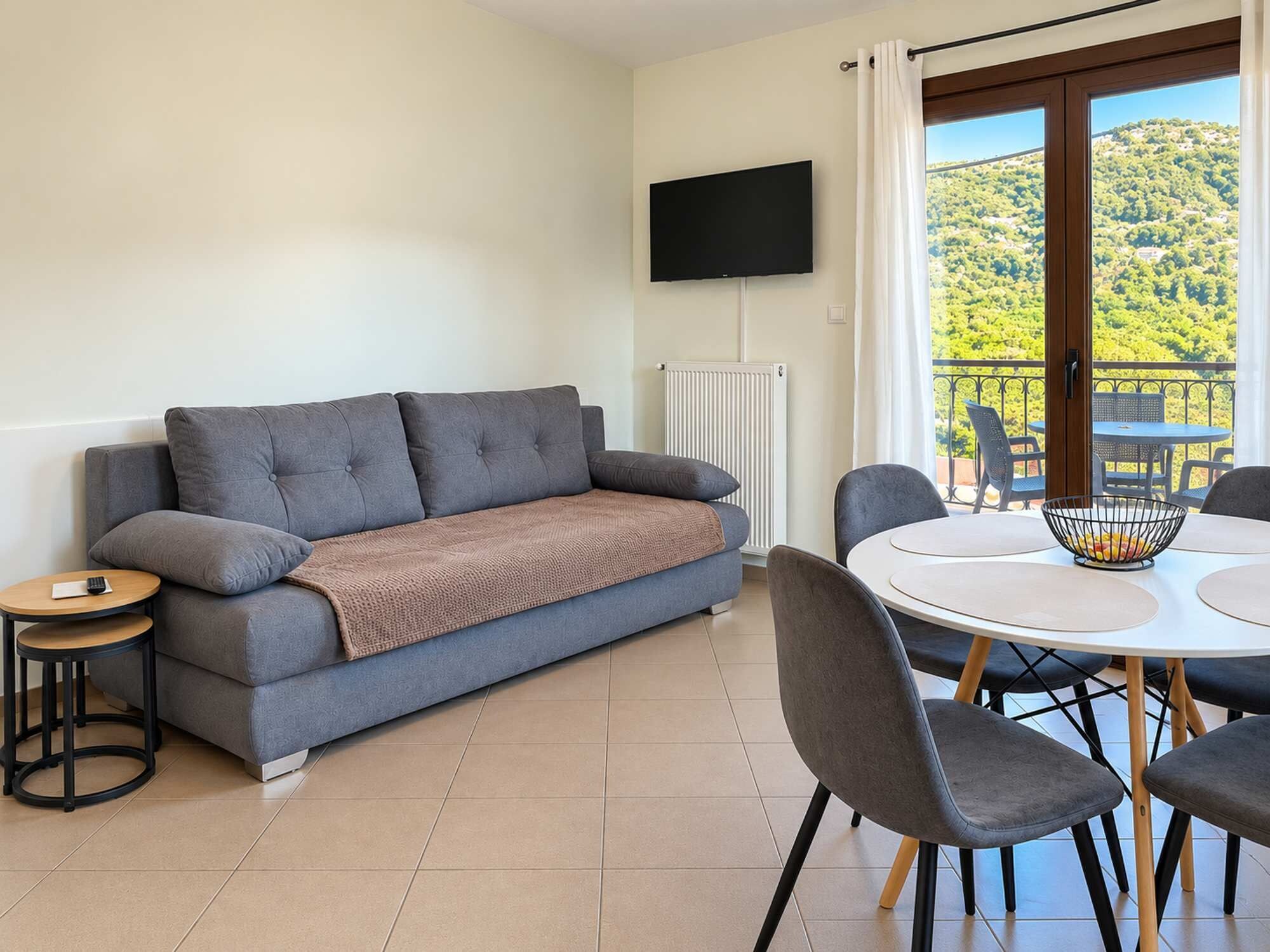 Ein modernes Wohnzimmer mit einer Couch, einem Fernseher, einem Esstisch mit Stühlen und Blick auf eine grüne Landschaft durch ein Fenster.