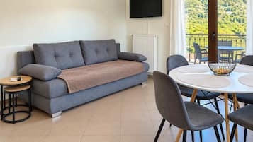 Ein modernes Wohnzimmer mit einer Couch, einem Fernseher, einem Esstisch mit Stühlen und Blick auf eine grüne Landschaft durch ein Fenster.