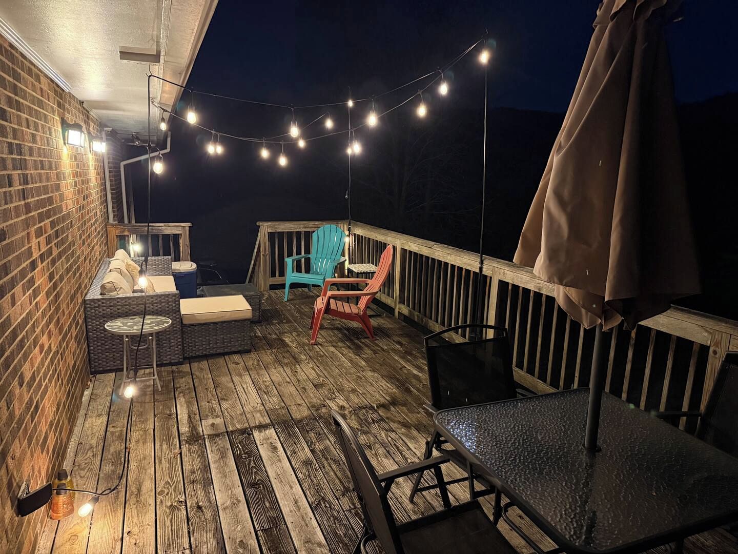 Een houten terras met buitenmeubilair, lichtjes en een bakstenen muur.