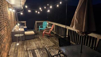 Een houten terras met buitenmeubilair, lichtjes en een bakstenen muur.