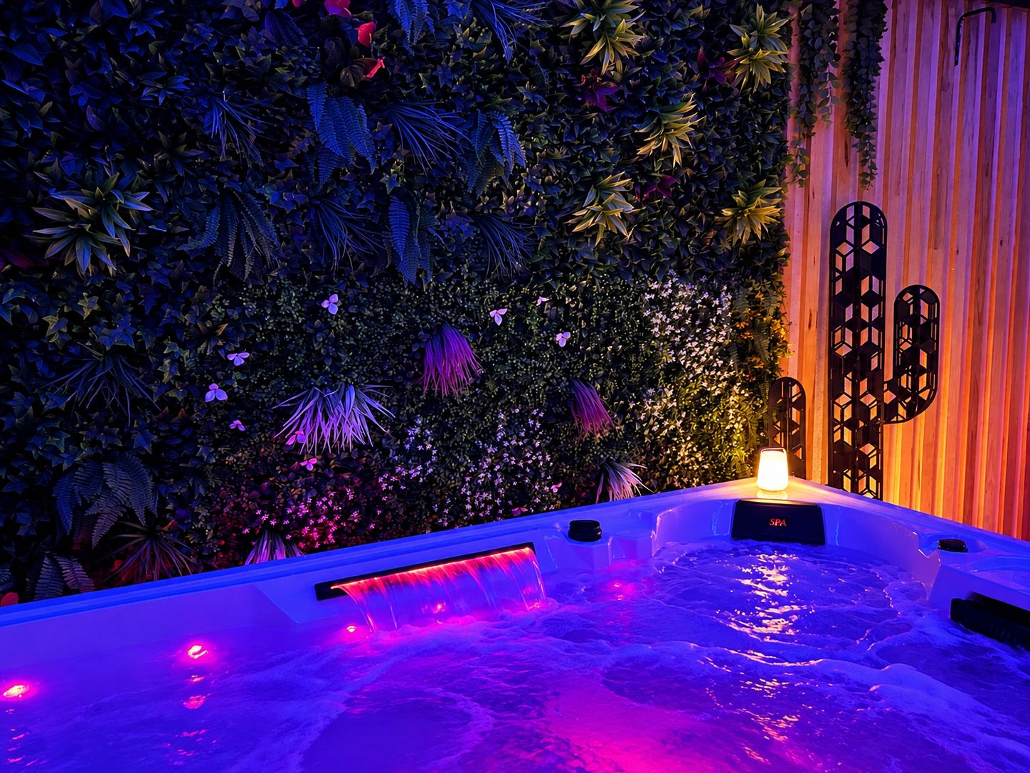 A hot tub with a waterfall feature surrounded by lush greenery and a wooden wall.